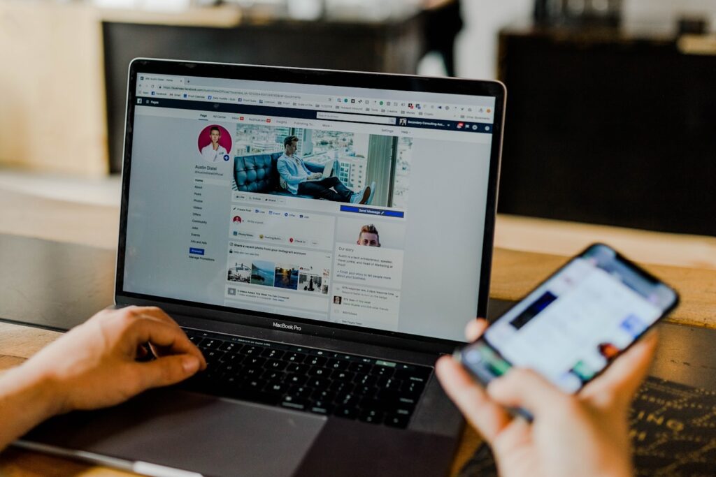 Persona che lavora a un portatile aperto su una pagina Facebook, con uno smartphone nell altra mano