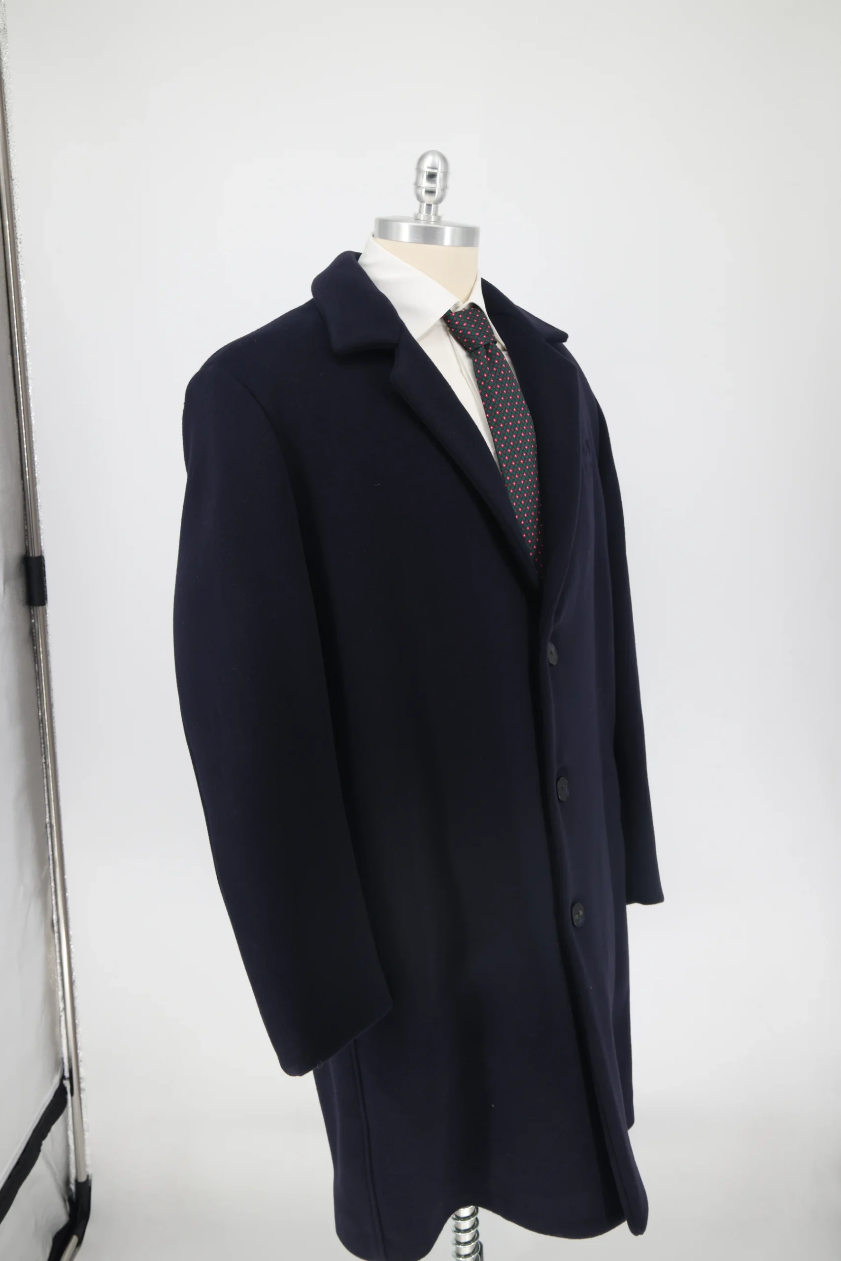 Scatto grezzo non ritoccato: cappotto blu su manichino davanti al fondale, con stativo metallico, pinza nera e base del treppiede in vista